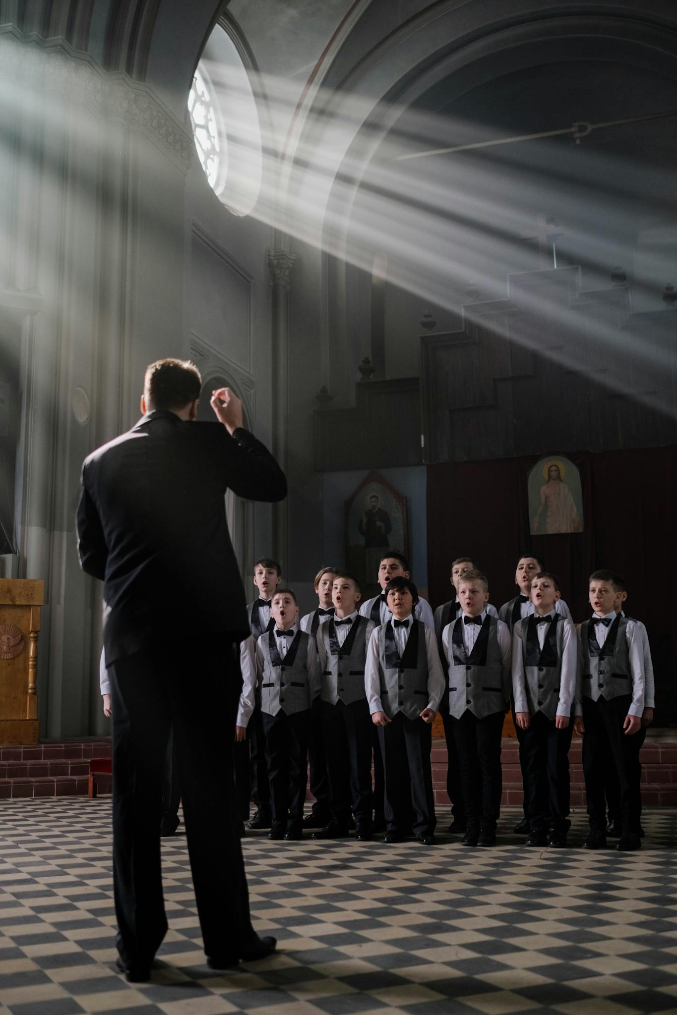 A boys choir rehearsing in a church, led by a conductor, under dramatic light beams.