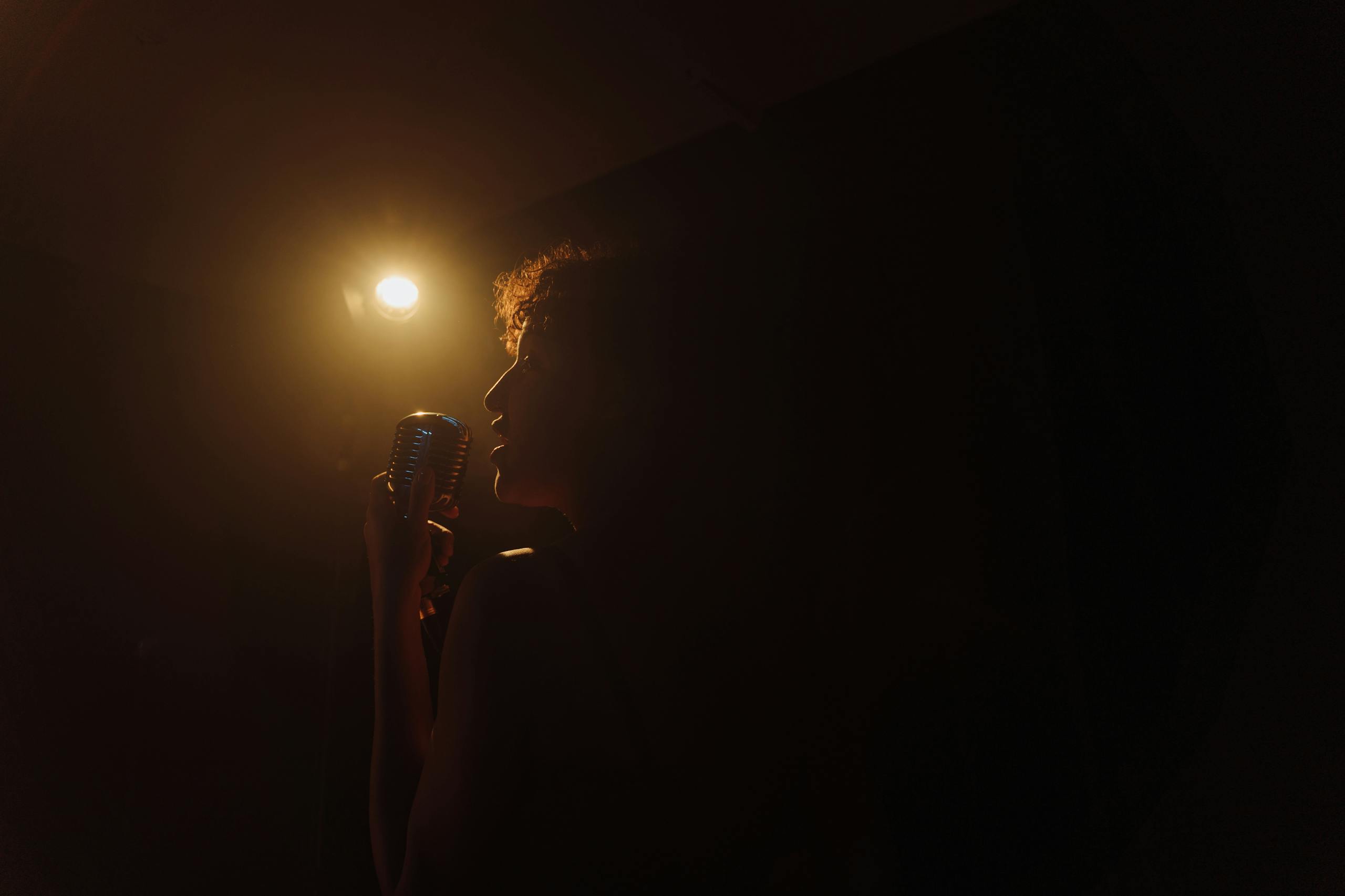 Vocalflow bietet professionelles Vocal Coaching für jeden Stimmtyp und fast jedes Genre an. A dramatic silhouette of a woman holding a vintage microphone, caught in warm stage light.