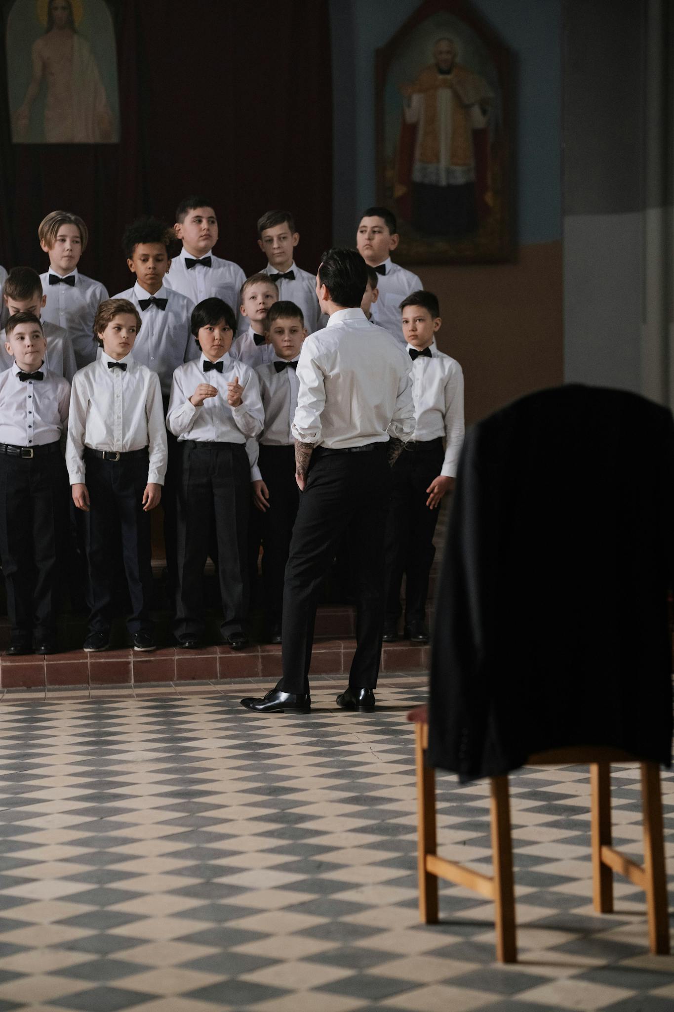 A group of boys in uniform bowties practicing choral singing indoors led by a conductor.