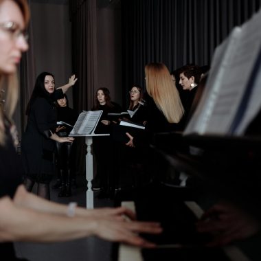 A group of female singers rehearsing with a conductor and pianist, focusing on music sheets.