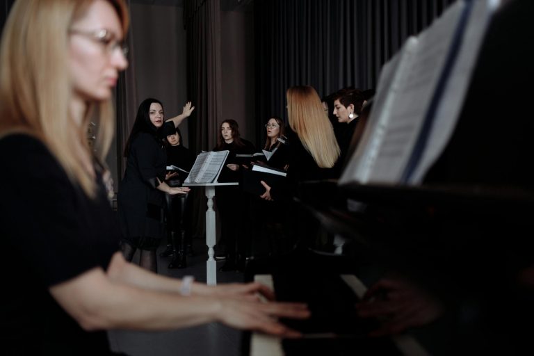 A group of female singers rehearsing with a conductor and pianist, focusing on music sheets.