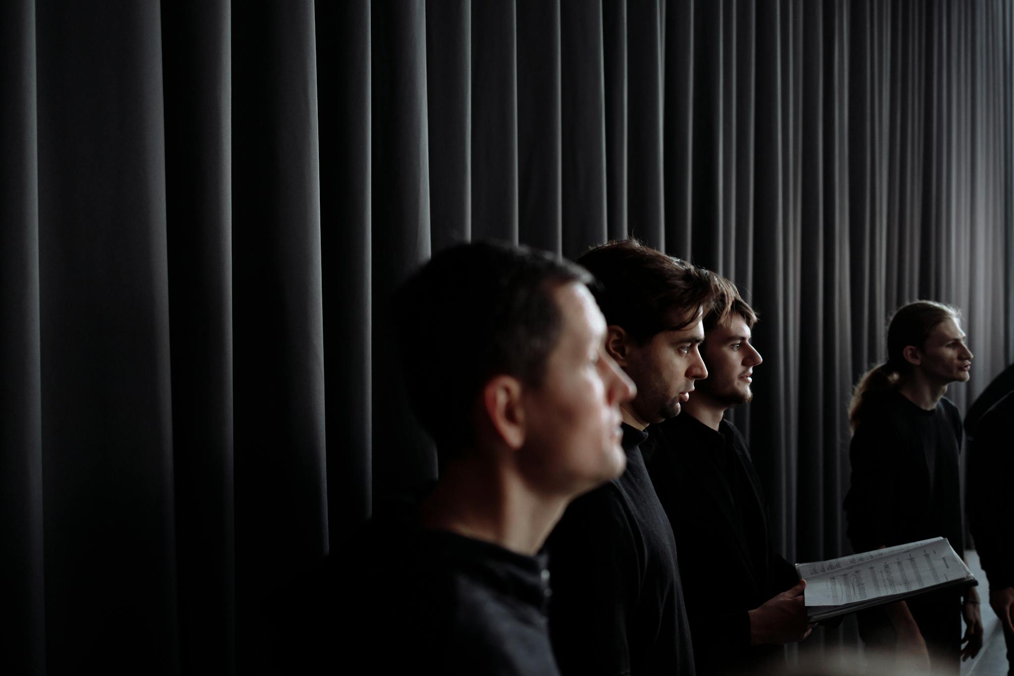 A group of male singers performing in a choir on a dark stage with moody lighting.