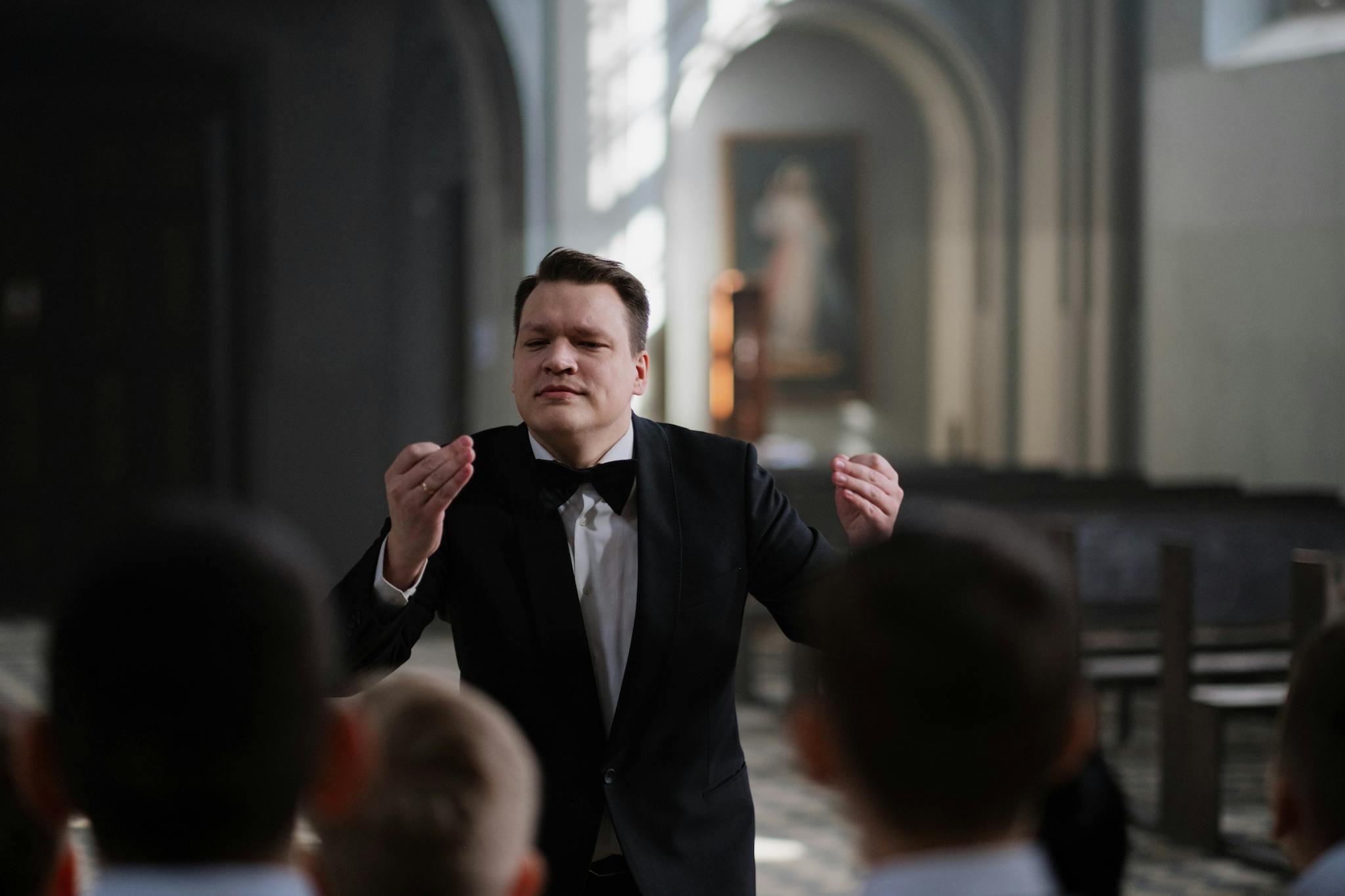 A male conductor leads a boys' choir rehearsal in a serene church setting.