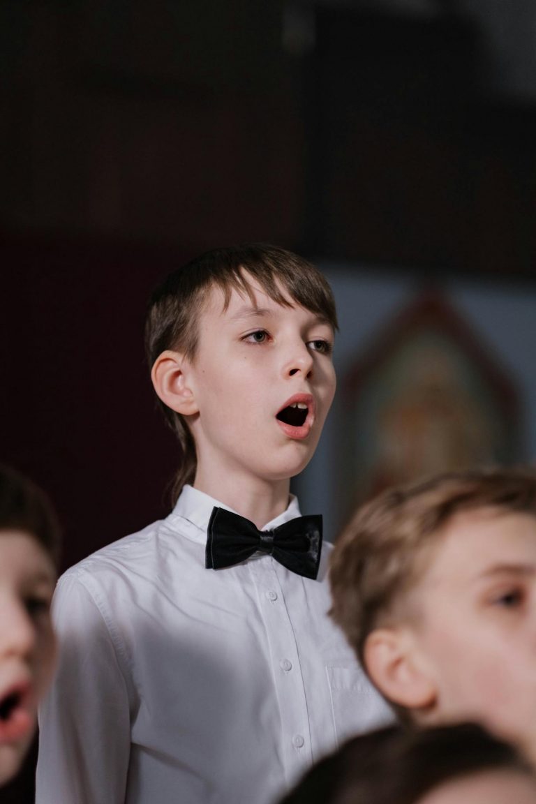 Boy in white shirt and bowtie singing in a choir practice session indoors.